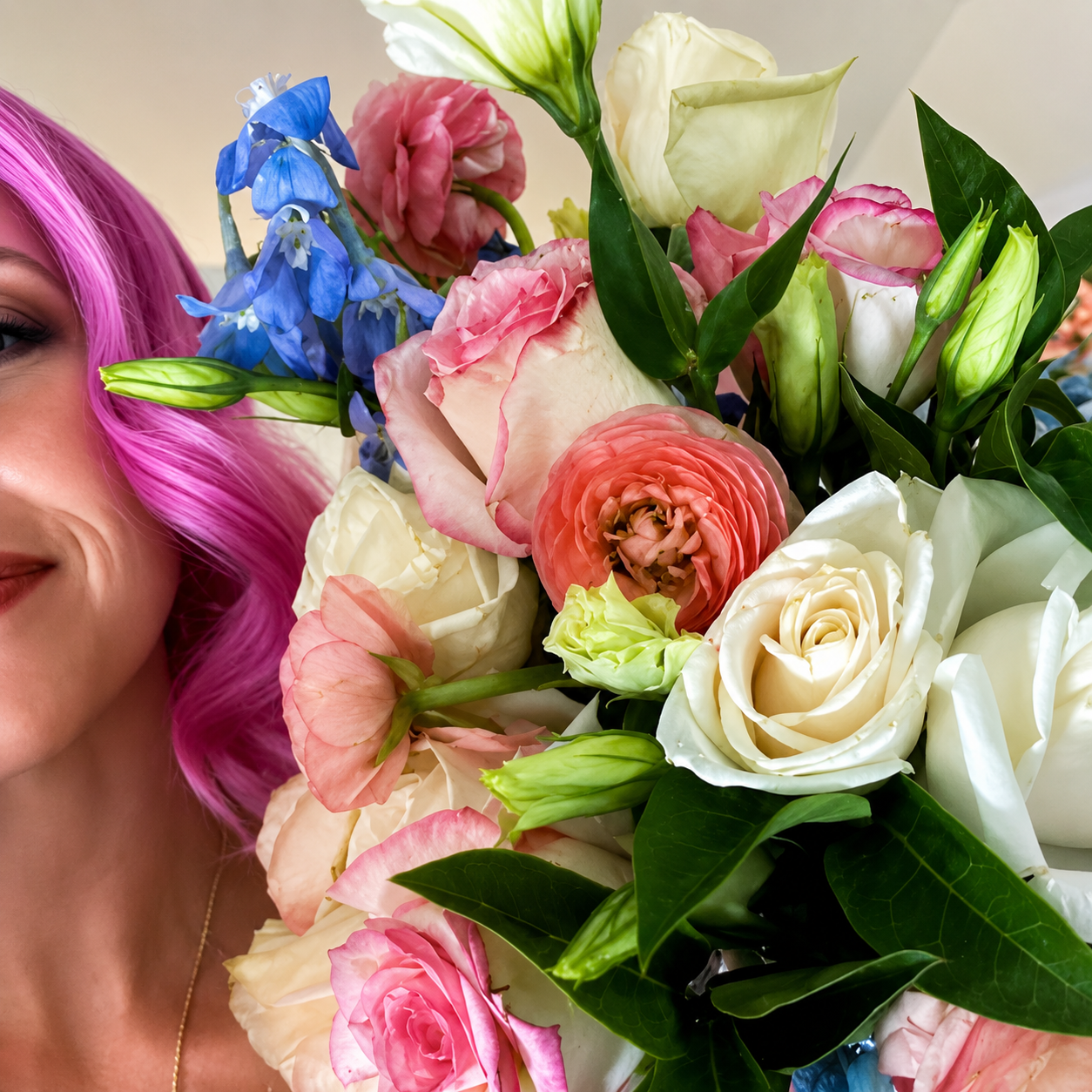 A woman with bright pink hair holding a lush bouquet of pink, white, and coral roses mixed with blue delphinium