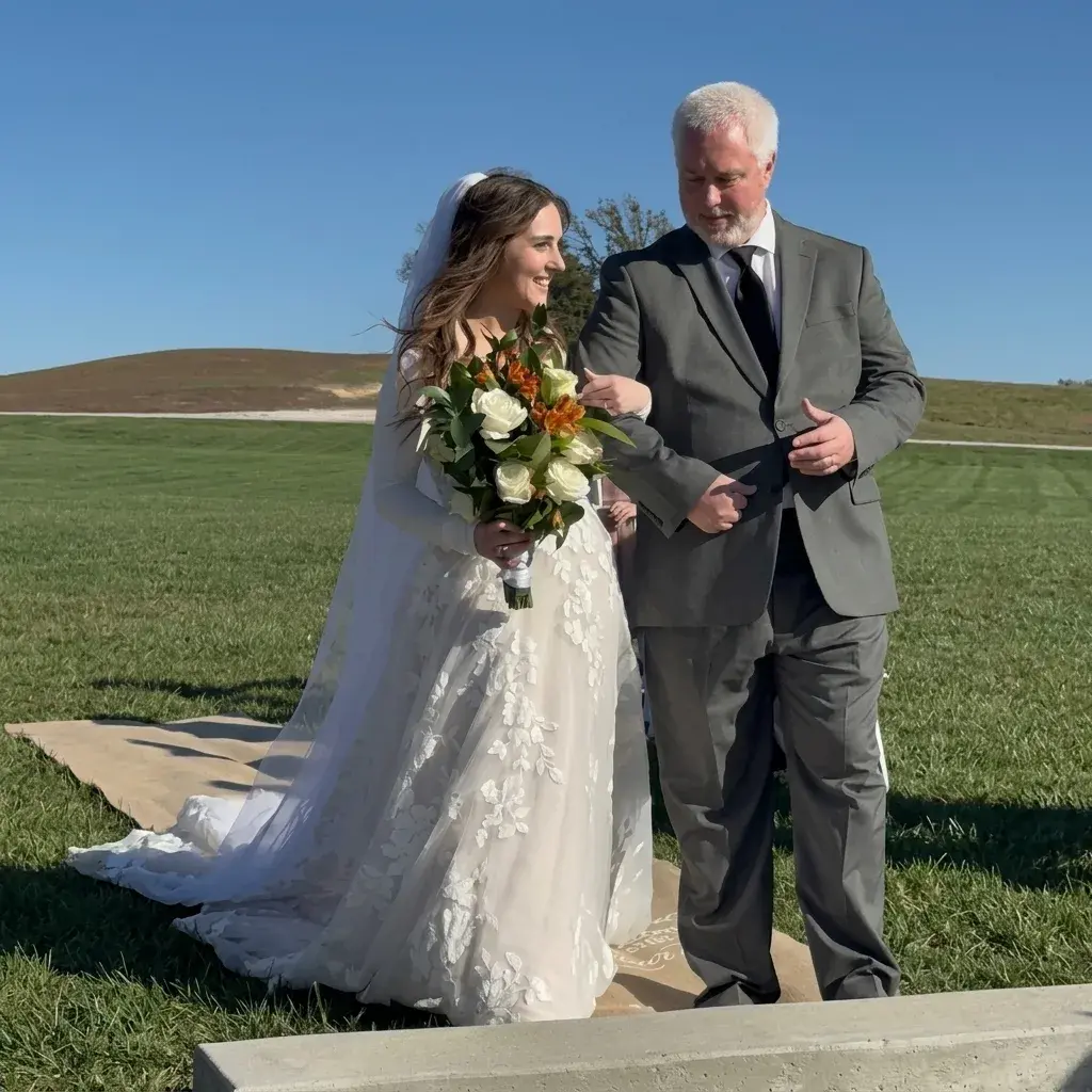 A smiling bride in a long-sleeved floral lace gown and veil, walking outdoors while holding a bouquet of white roses and orange alstroemeria.